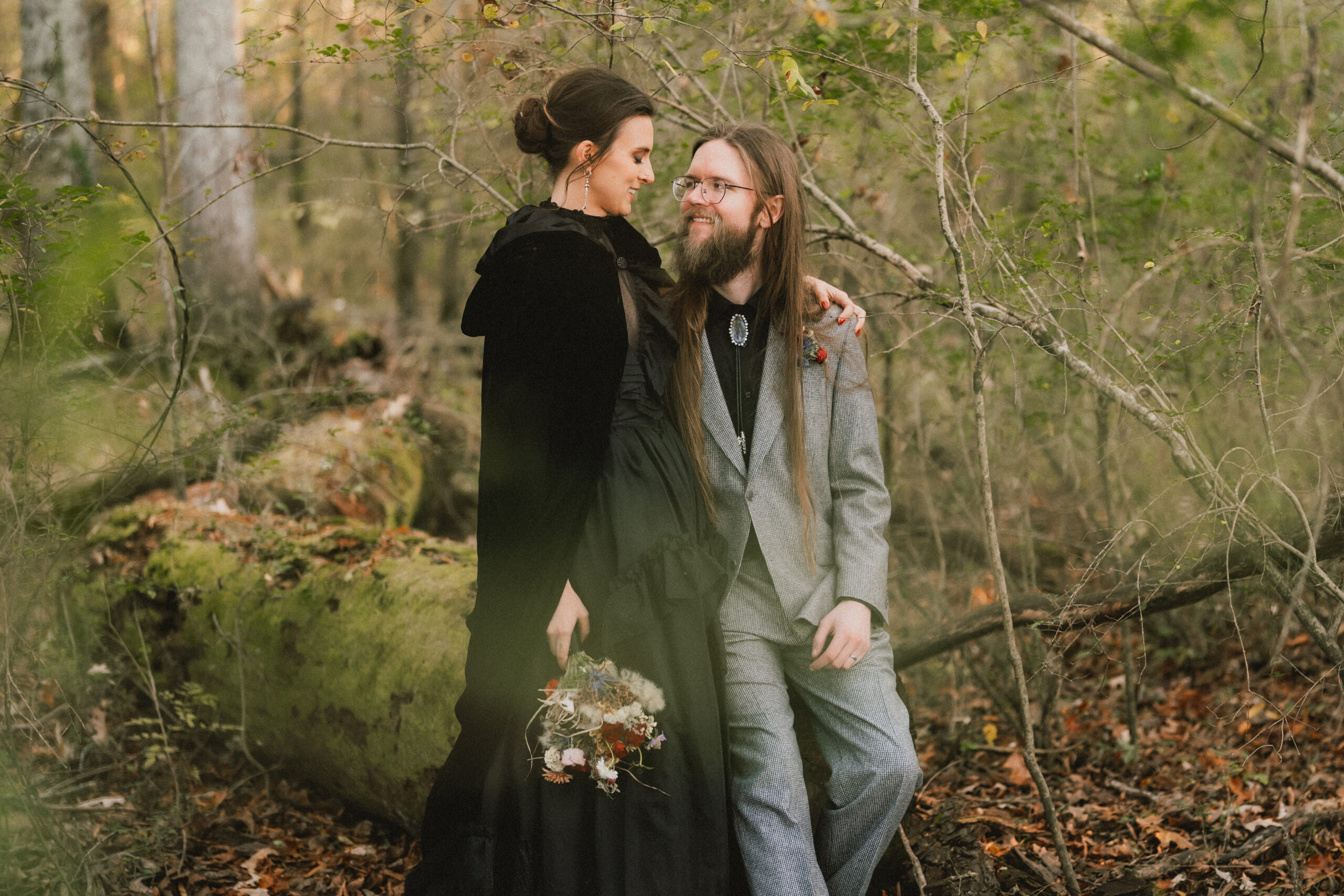 Erin & Baxter smile by a mossy log in the woods, her black cloak and dried bouquet at Covey Creek
