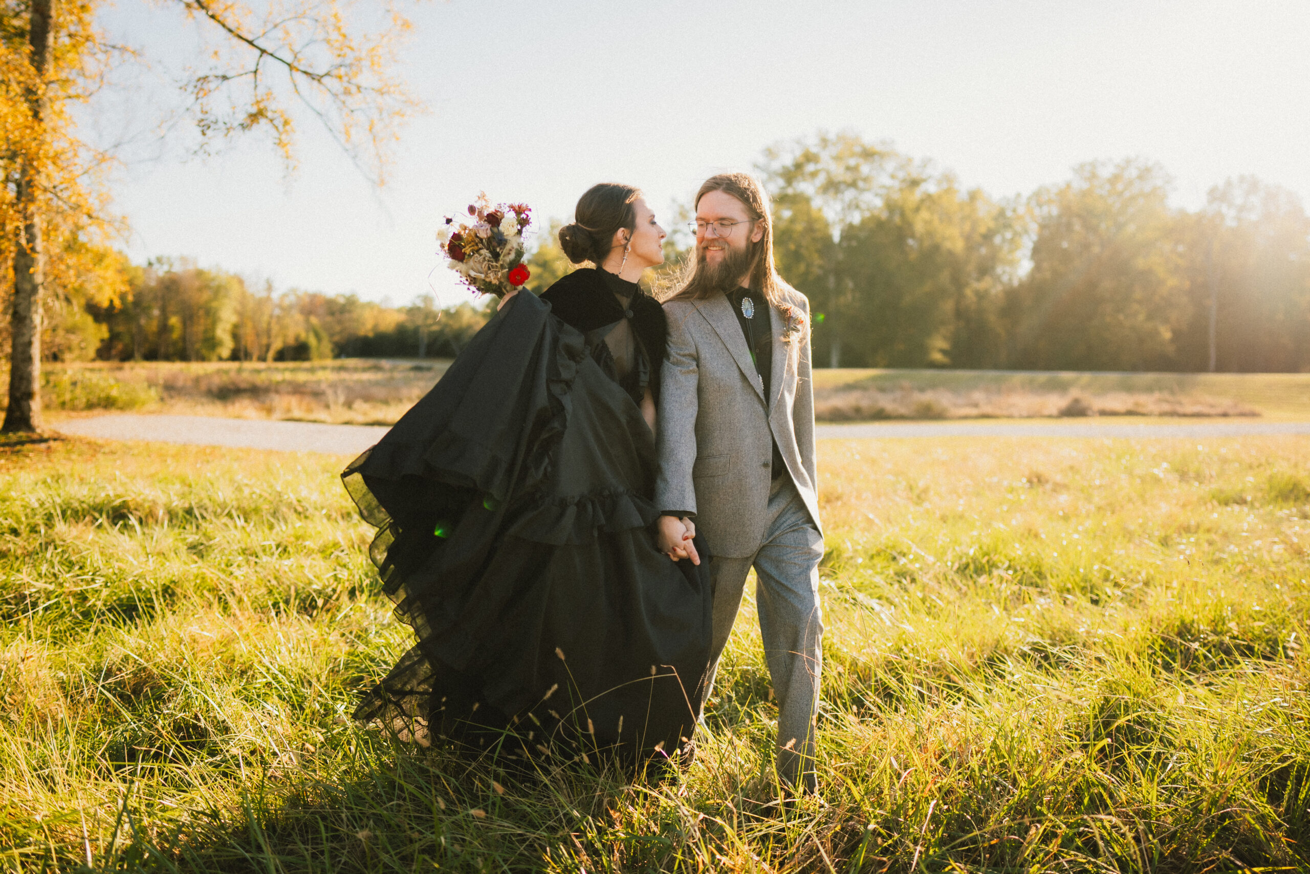 Erin & Baxter walk hand-in-hand through sunlit autumn grass in black gown and grey suit at Covey Creek