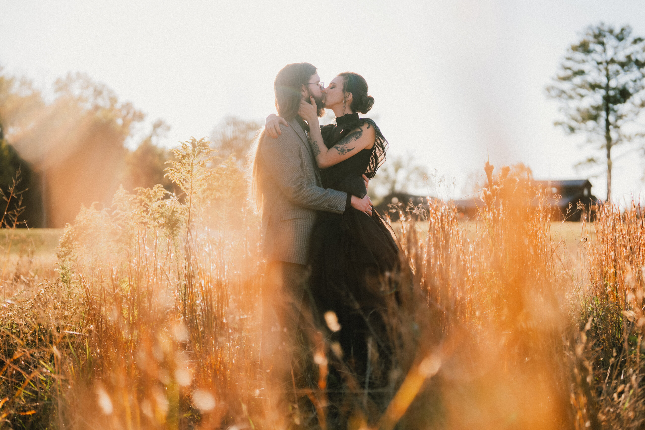 Erin & Baxter share a kiss in tall golden grass at sunset, barn glowing behind them at Covey Creek