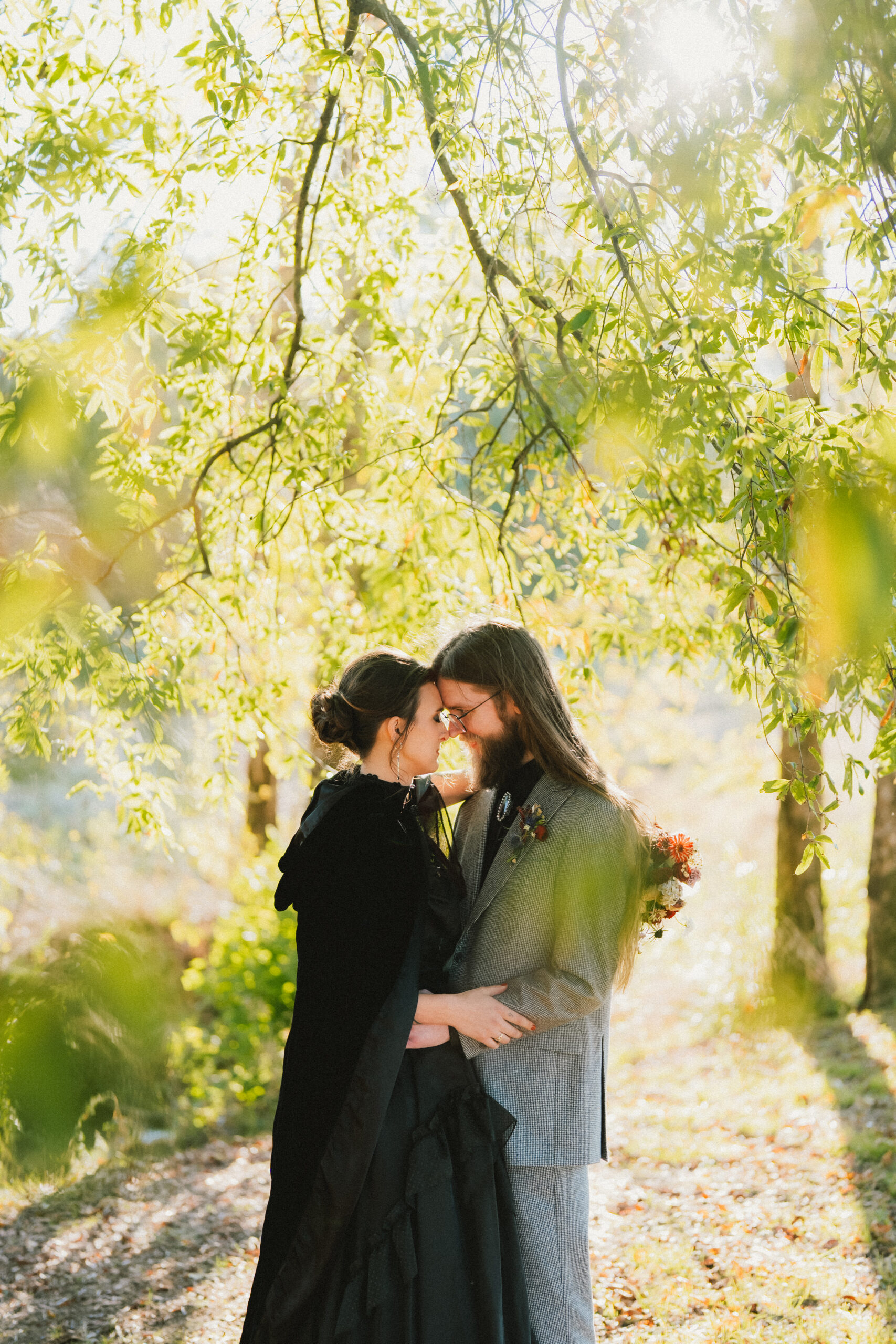Erin & Baxter touch foreheads under sunlit trees, her black velvet cloak and grey suit at Covey Creek