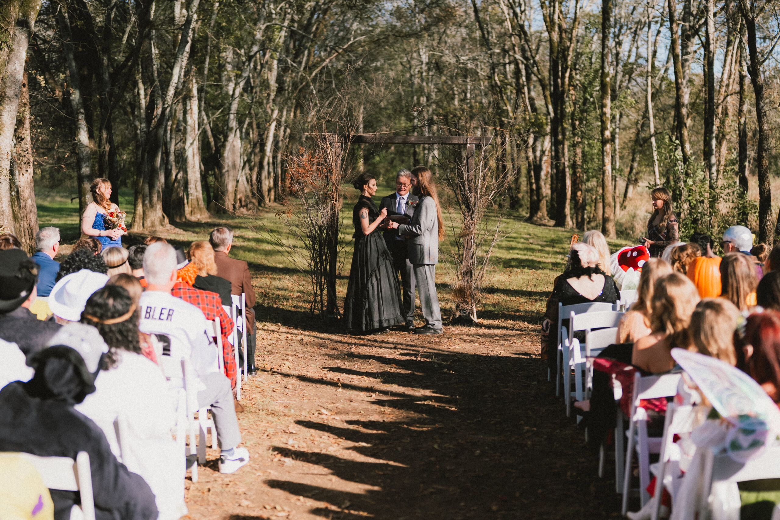 Erin & Baxter exchange vows under a wooden branch arch as costumed guests watch at Covey Creek