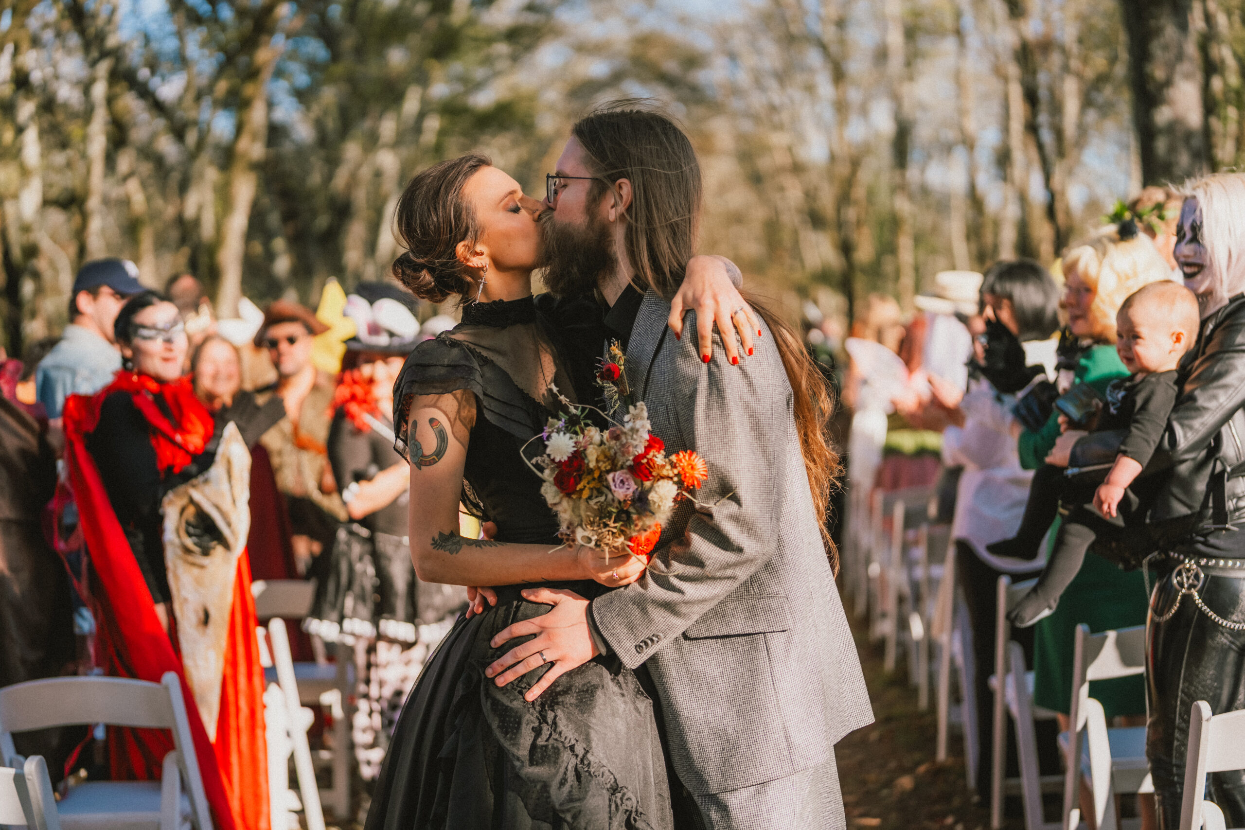Erin & Baxter kiss in the outdoor ceremony aisle among costumed guests under trees at Covey Creek