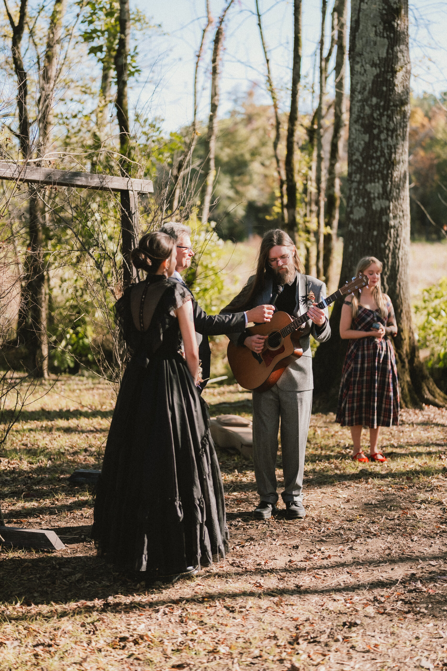 Erin & Baxter's ceremony: Baxter plays guitar for Erin under a wooden arch in woods at Covey Creek