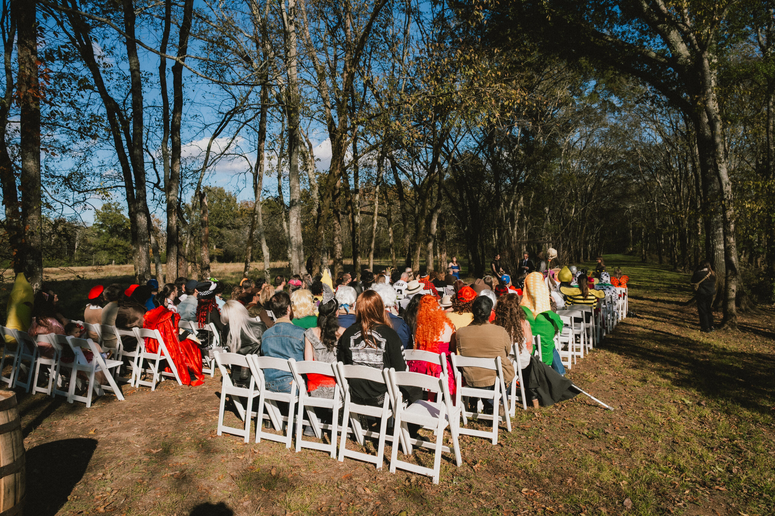Costumed guests in banana and dinosaur suits face Erin & Baxter's wooded ceremony aisle at Covey Creek