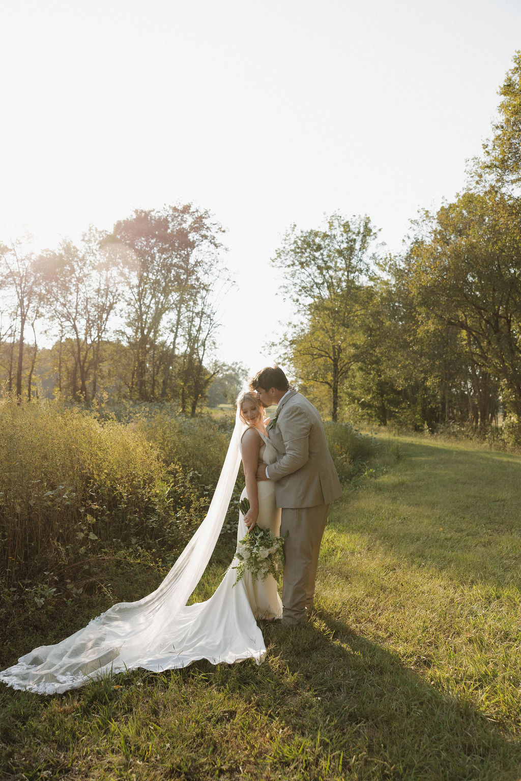 Joseph kisses Hailey's forehead in a sunlit grassy field during Hailey & Joseph portraits at Covey Creek