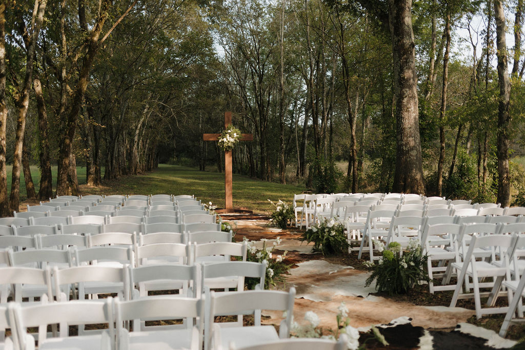 Empty white chairs and cowhide aisle lead to a flowered wooden cross at Covey Creek for Hailey & Joseph