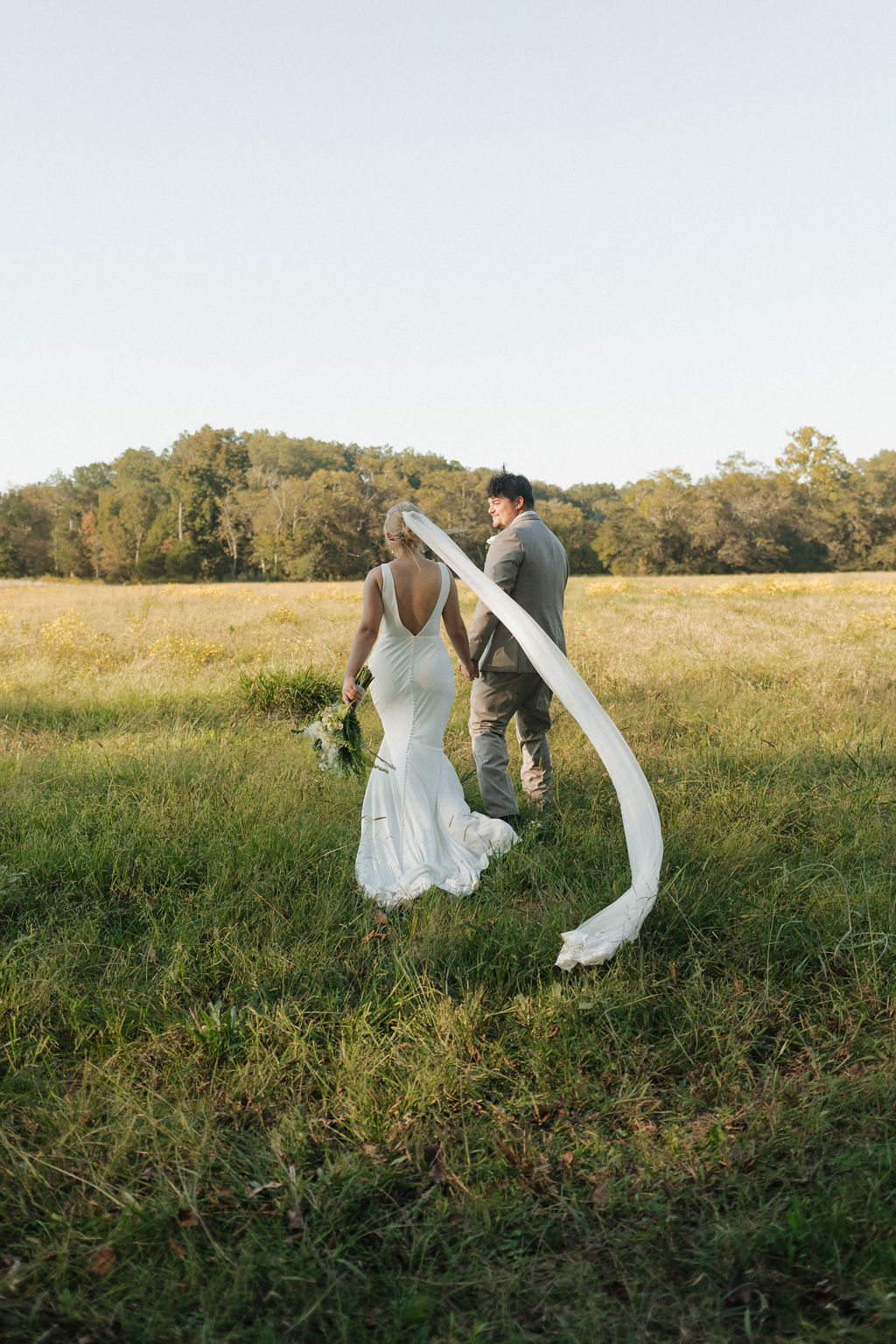 Hailey & Joseph walk hand-in-hand through a wildflower field, her veil blowing, at Covey Creek