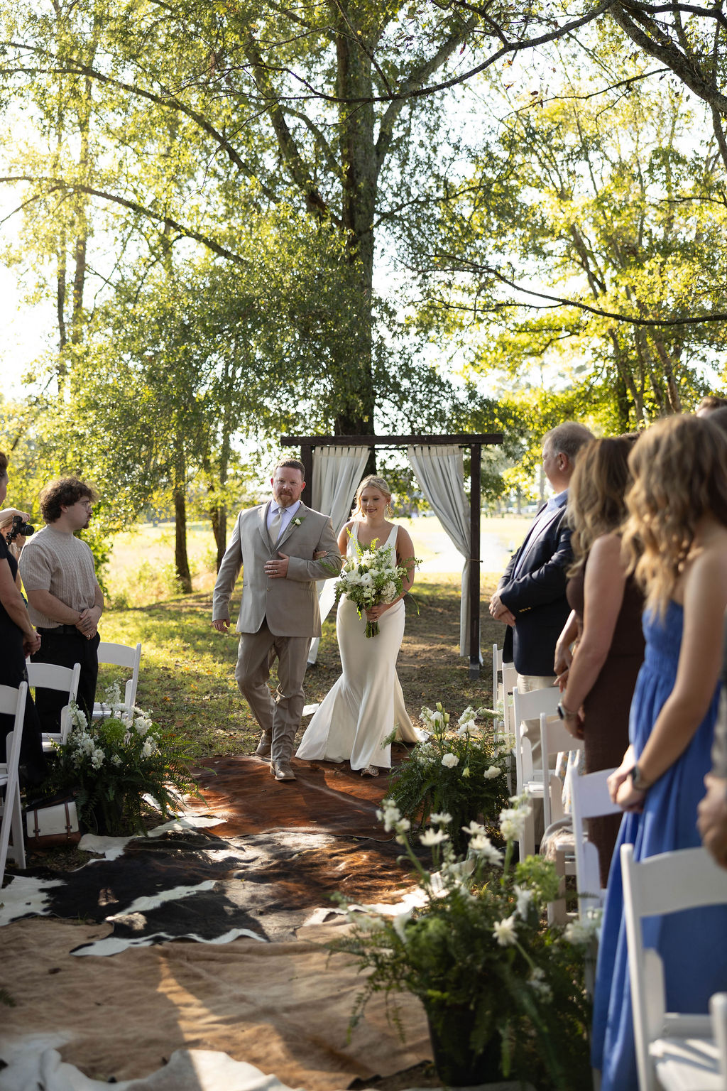 Hailey walks the cowhide aisle with her father past white florals at Hailey & Joseph's Covey Creek ceremony