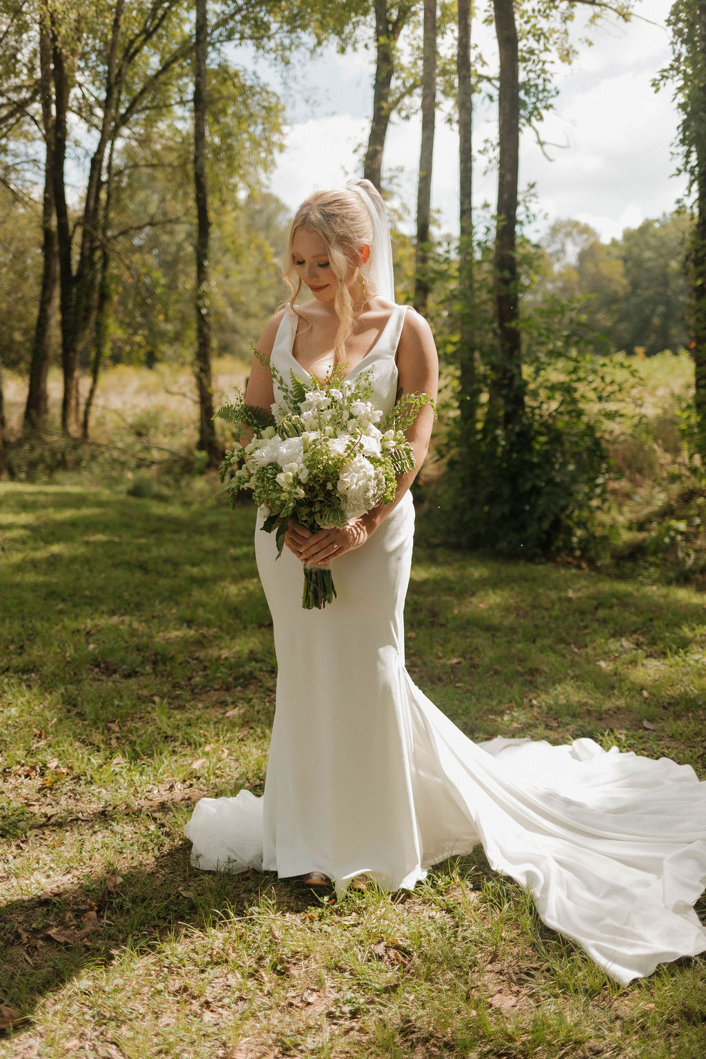 Hailey looks down at her white and green bouquet in a wooded clearing at Covey Creek for Hailey & Joseph