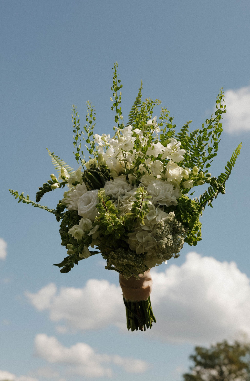 White rose and fern bridal bouquet held against a blue sky at Hailey & Joseph's wedding at Covey Creek