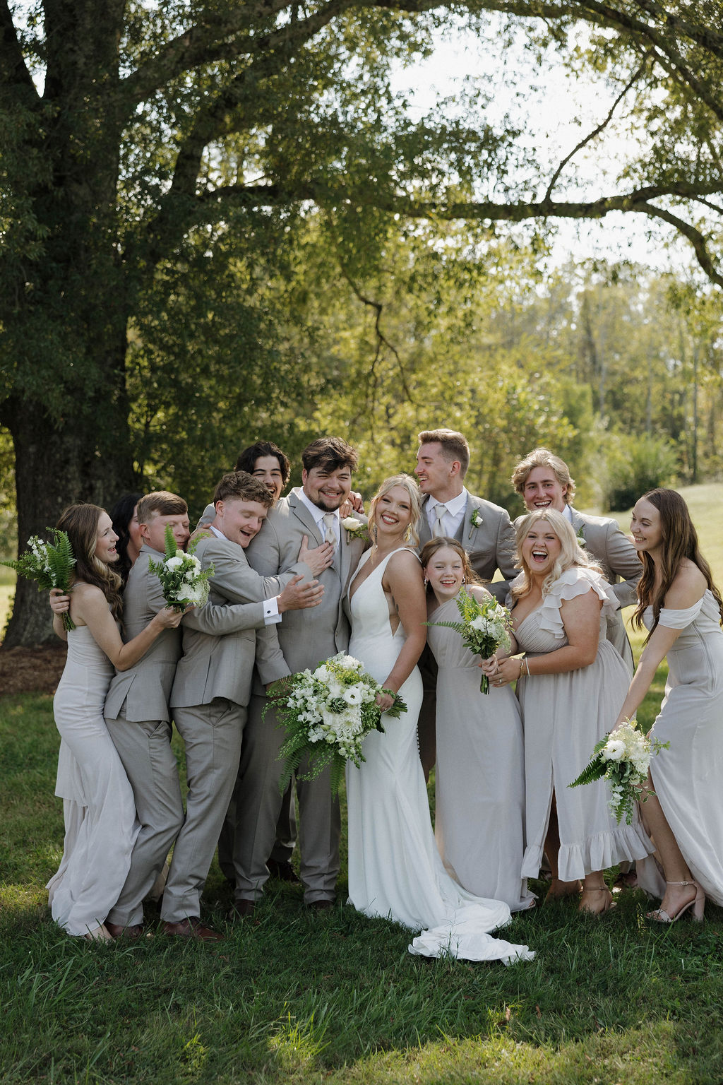 Hailey & Joseph laugh with their wedding party under a shade tree on the lawn at Covey Creek