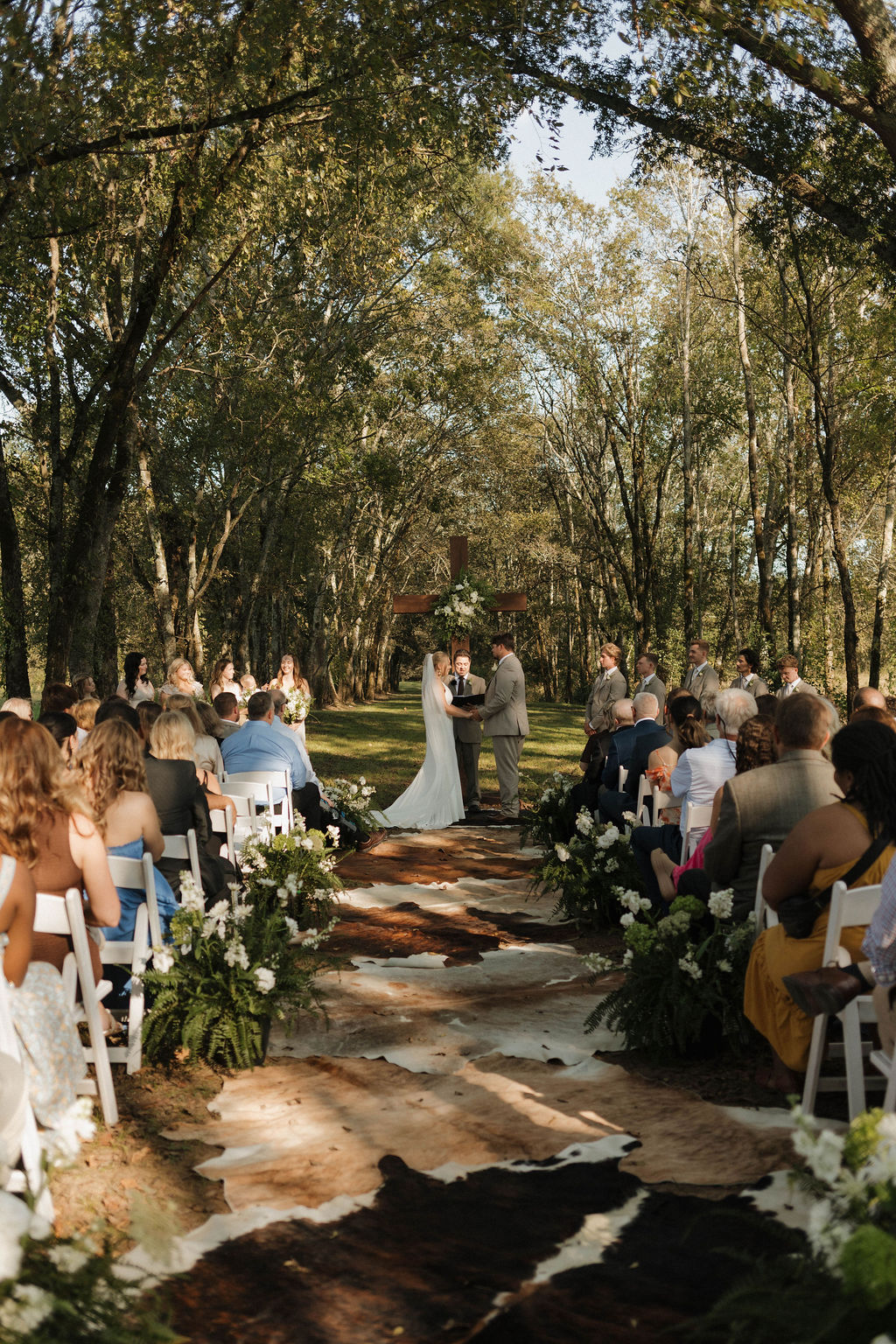 Guests face Hailey & Joseph at the wooden cross along a cowhide aisle under Covey Creek trees