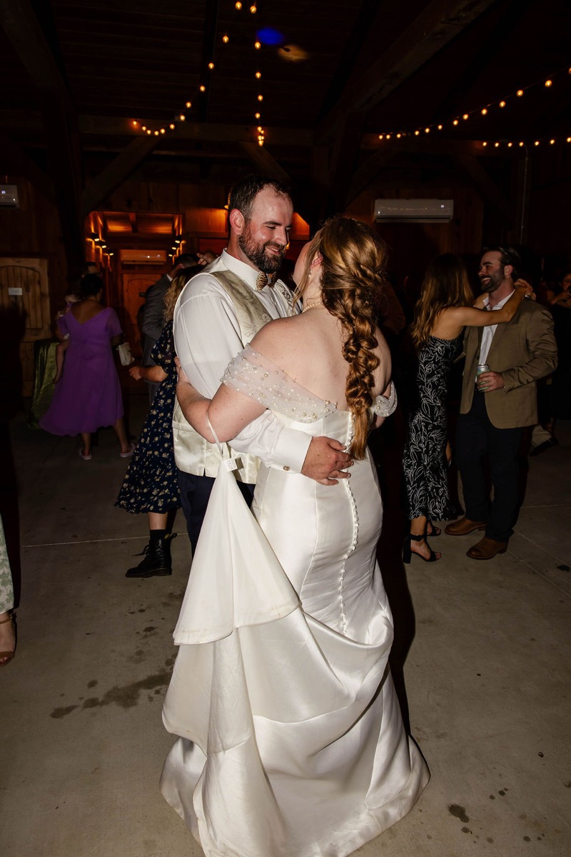 Hannah & Nicholas slow dance in the Covey Creek barn, her braid and button-back gown lit by string lights.