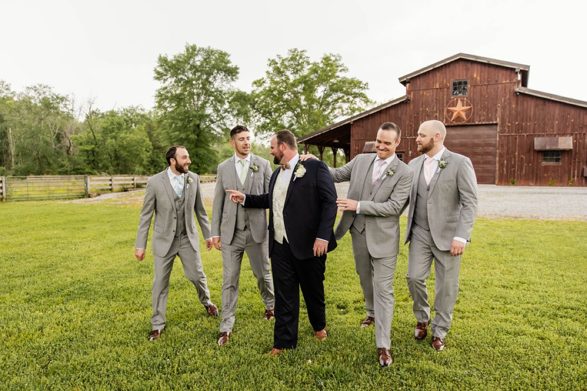 Nicholas in navy and grey-suited groomsmen laugh crossing the lawn before Covey Creek's barn, Hannah & Nicholas.