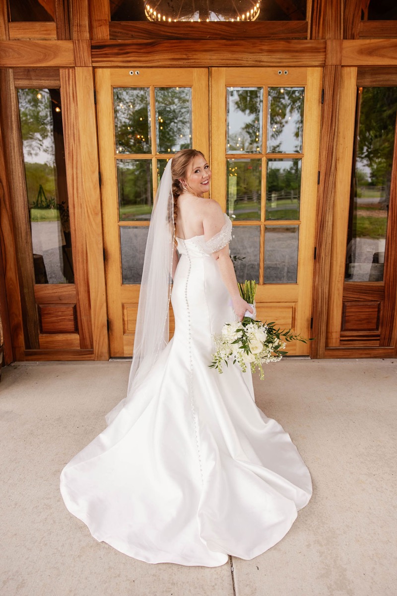 Hannah looks back over her shoulder in a button-back gown and veil by Covey Creek's glass doors, Hannah & Nicholas.