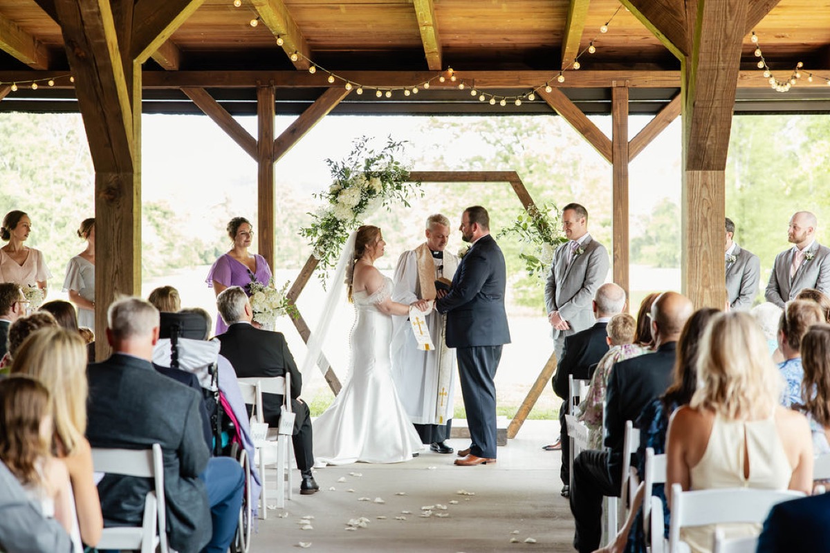 Hannah & Nicholas hold hands under a white-flower hex arch as guests watch at Covey Creek.