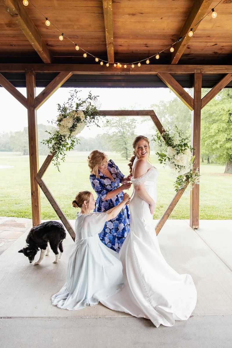 Hannah smiles in her button-back gown under Covey Creek's flowered hex arch as helpers zip her, Hannah & Nicholas.