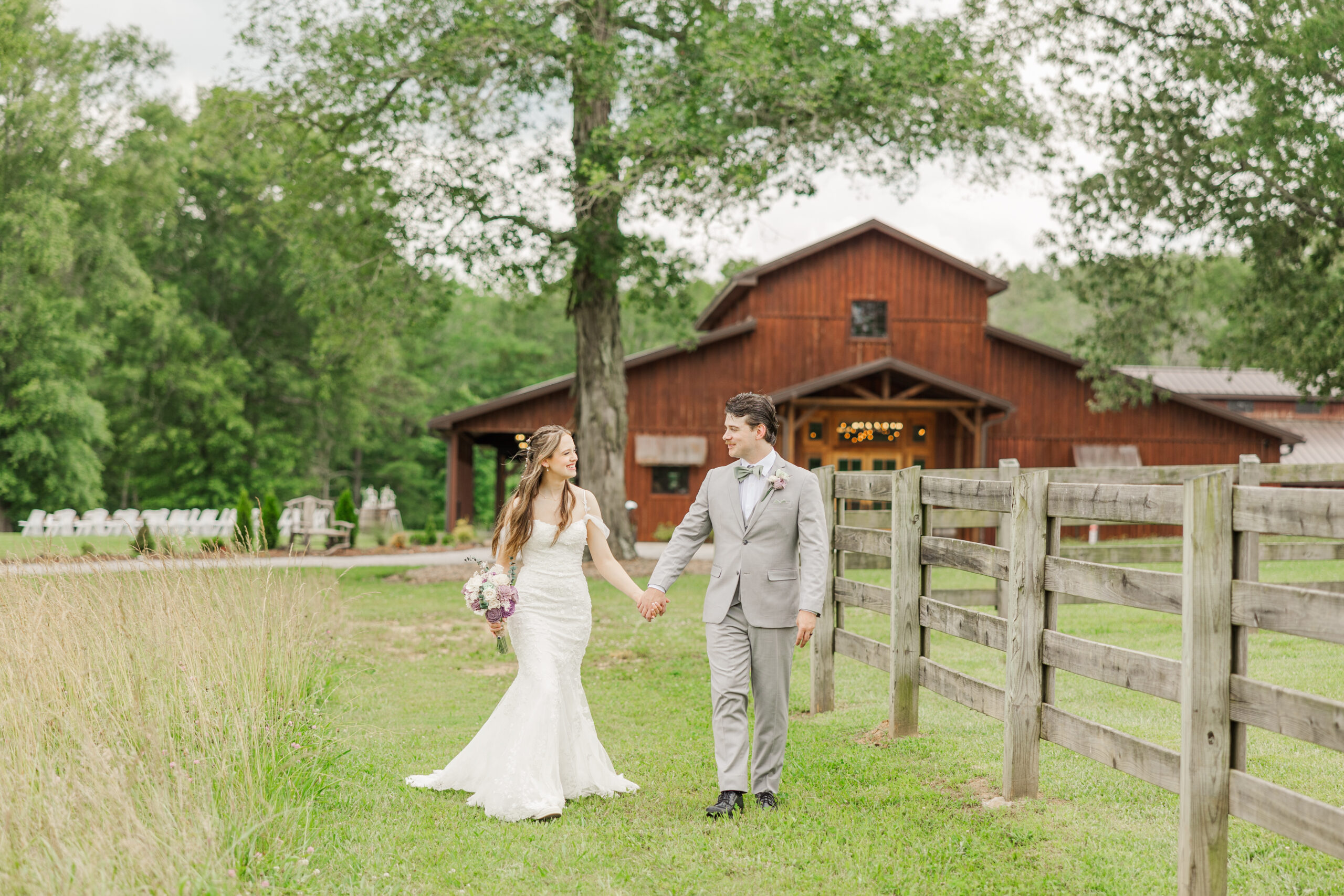 Hannah & Ryan stroll past a wooden fence by the barn, ceremony chairs beyond, at Covey Creek.