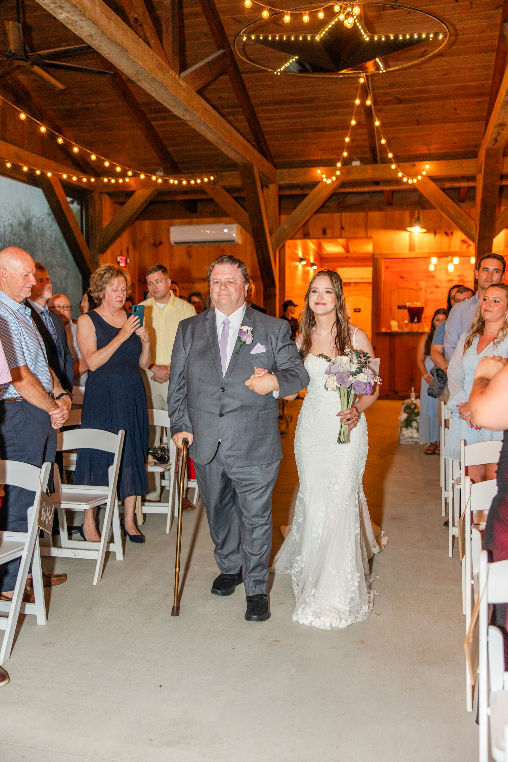 Hannah walks the barn aisle on her father's arm toward Ryan under string lights at Covey Creek.