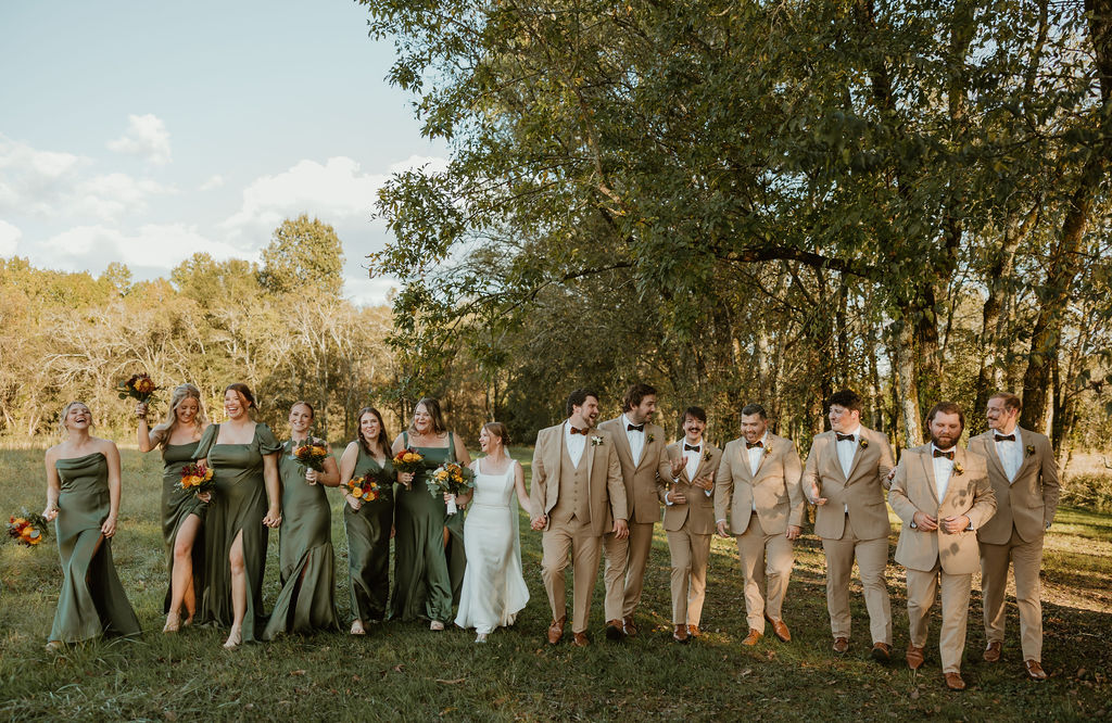 JJ & Will lead olive bridesmaids and tan groomsmen walking through a sunny field at Covey Creek