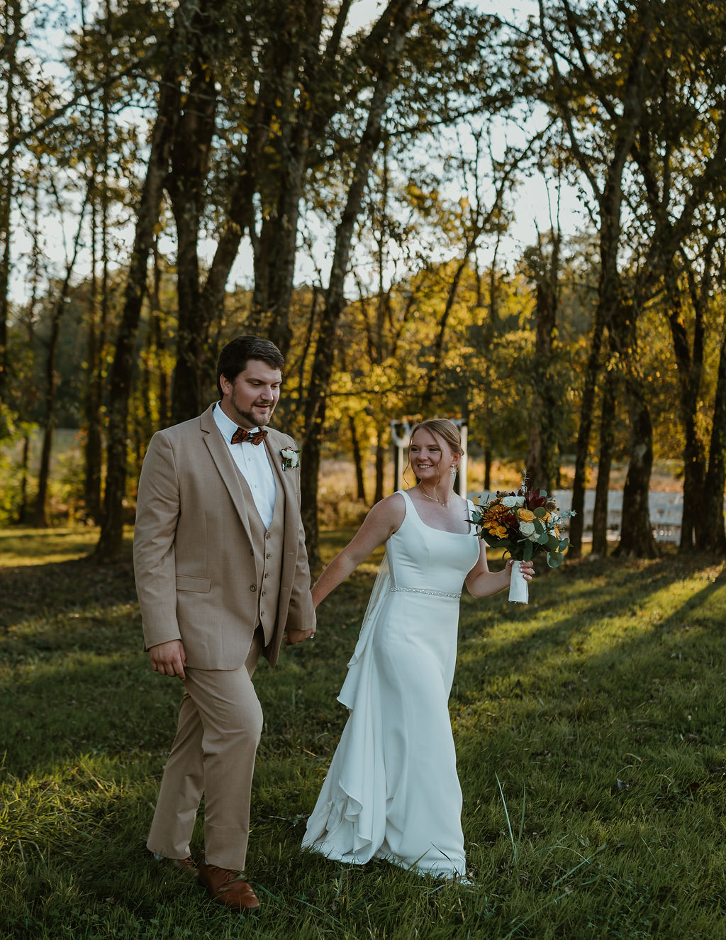 JJ & Will walk hand in hand across the field in golden light, ceremony trees behind at Covey Creek
