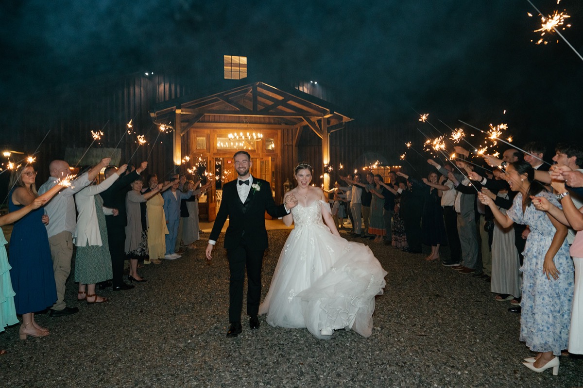 Katelyn & Josh walk a sparkler exit at night past Covey Creek’s lit barn entrance with cheering guests
