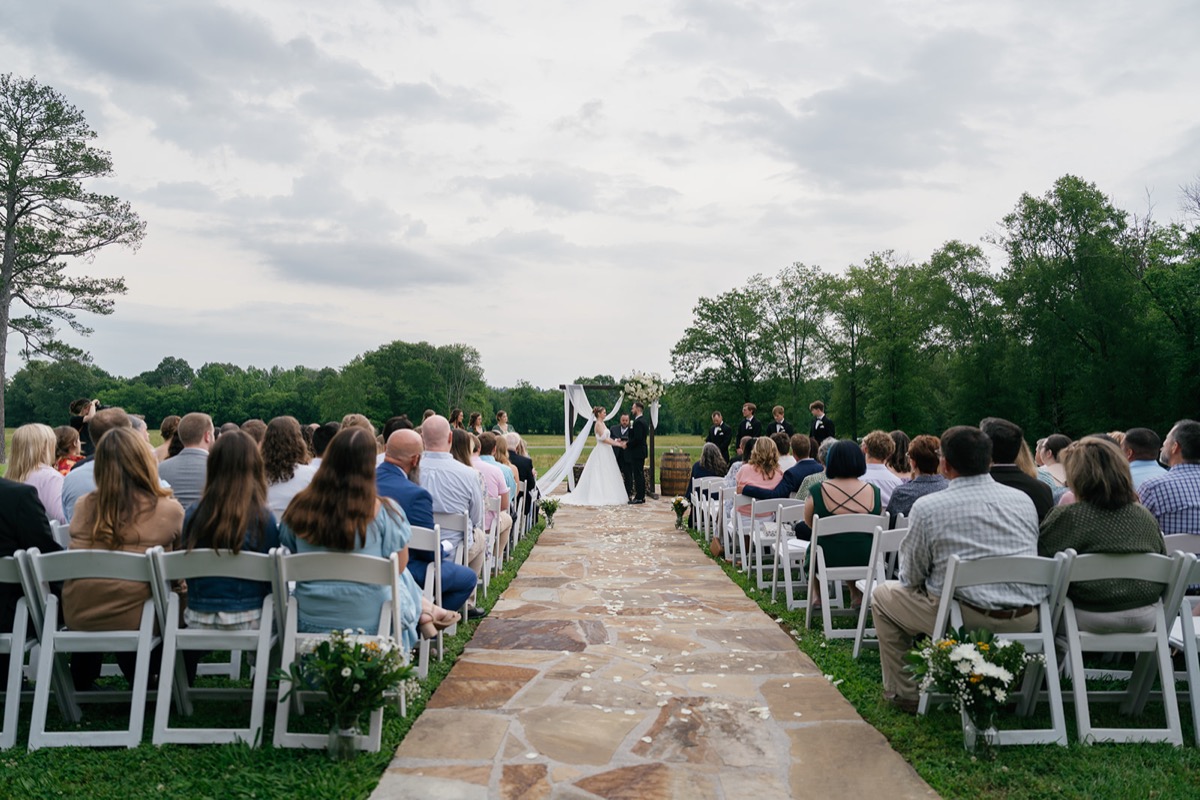 Katelyn & Josh stand at Covey Creek’s Patio arbor as guests watch from white chairs along the stone aisle