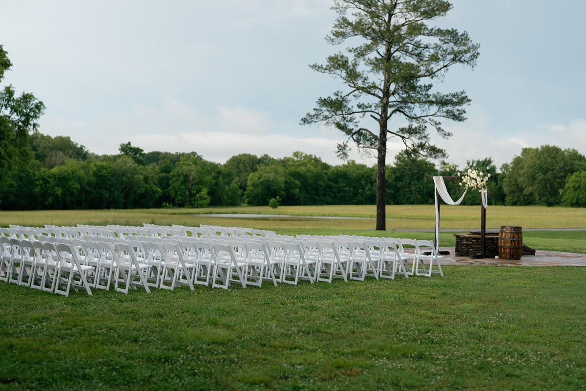 Katelyn & Josh Patio ceremony setup at Covey Creek: white chairs facing a draped wooden arbor by the pond