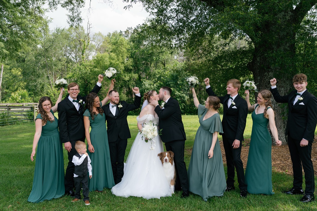 Katelyn & Josh kiss as their Covey Creek wedding party cheers, dog at their feet on the green lawn