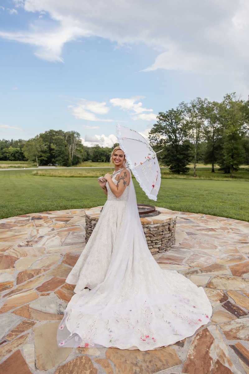 Kolbe smiles with a lace parasol and floral embroidered train on the firepit patio at Covey Creek, Kolbe & Nathan