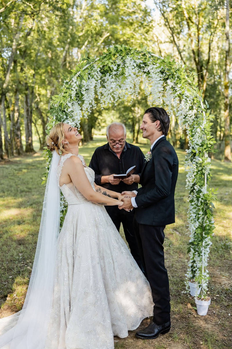 Kolbe laughs with Nathan under a white floral arch during their Tunnel of Love ceremony at Covey Creek