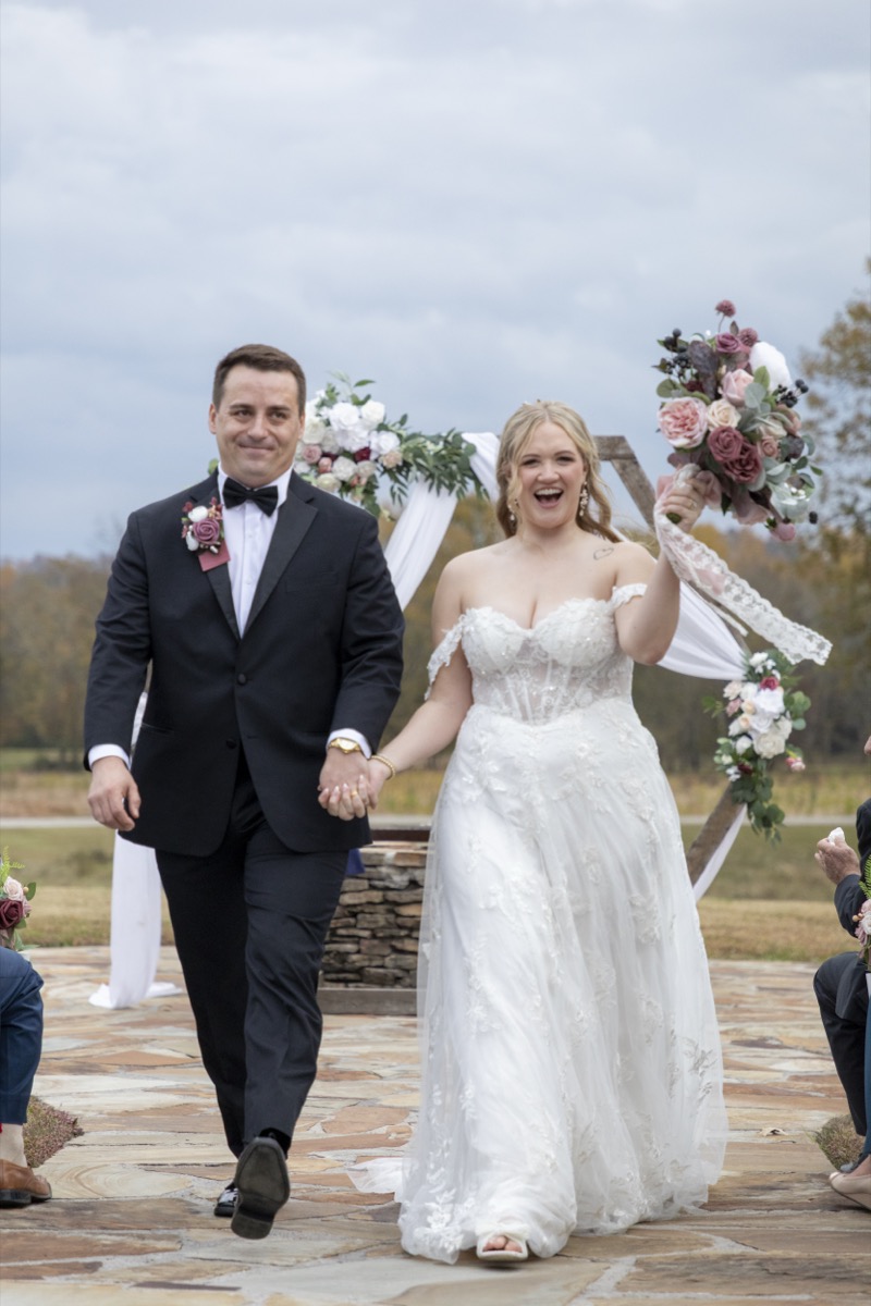 Kristen & Dalton smile hand-in-hand walking the aisle after vows, mauve bouquet trailing lace ribbon at Covey Creek