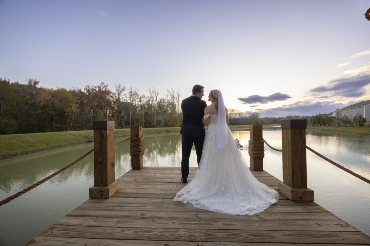 Kristen & Dalton stand arm-in-arm on the Pond dock overlooking calm water and fall trees at Covey Creek