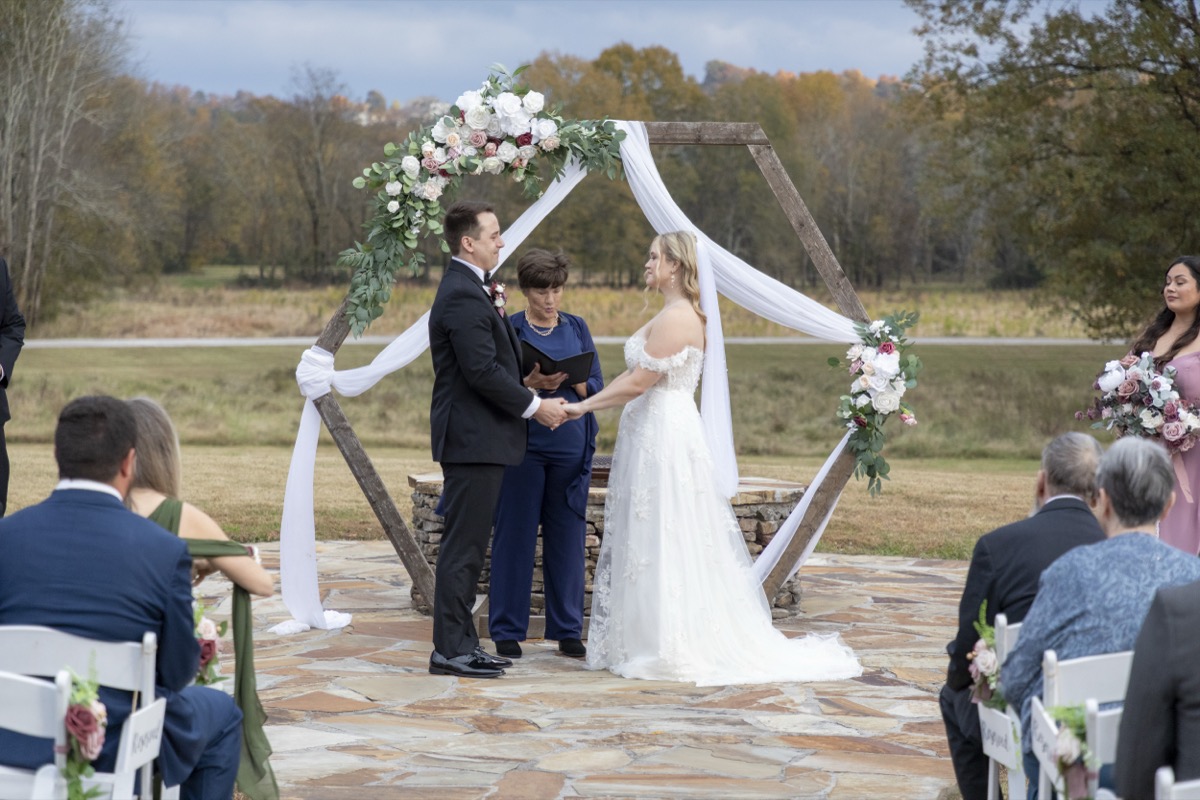 Kristen & Dalton hold hands under a floral hexagonal arbor with their officiant at the Firepit at Covey Creek