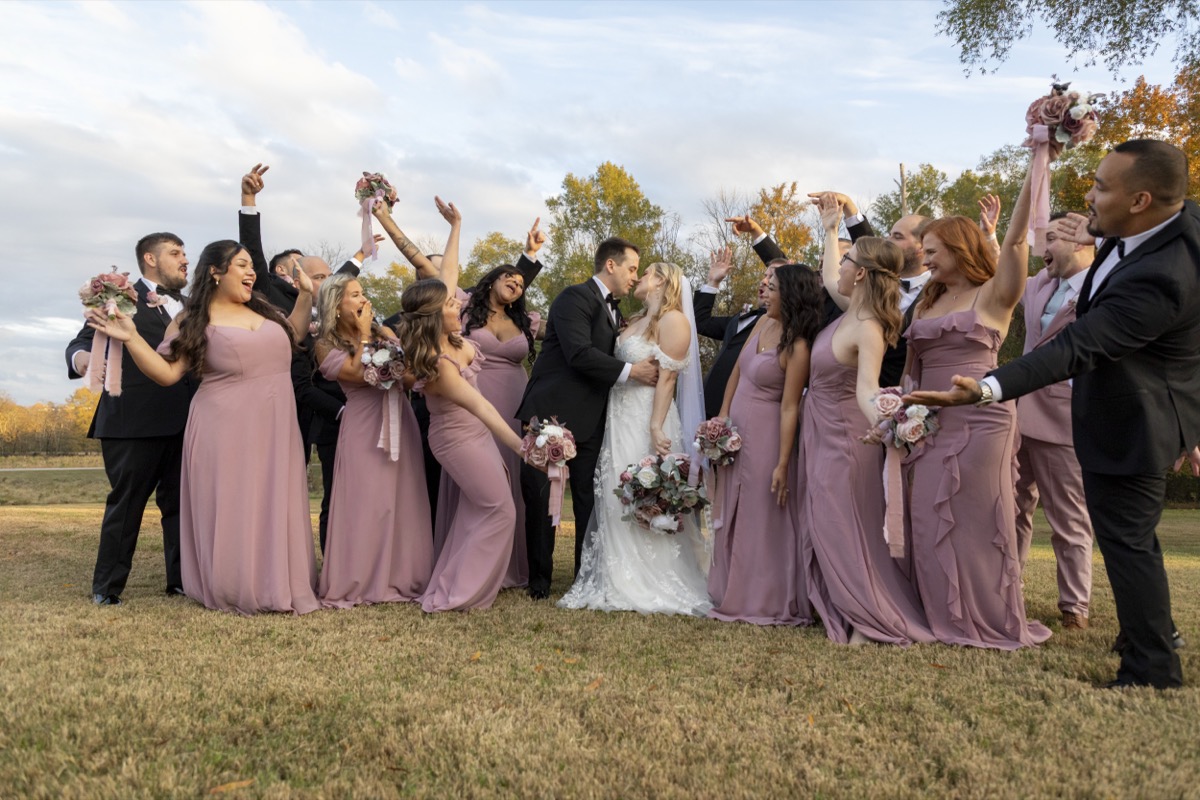 Kristen & Dalton kiss as their bridal party in mauve gowns and black tuxedos cheers in a field at Covey Creek