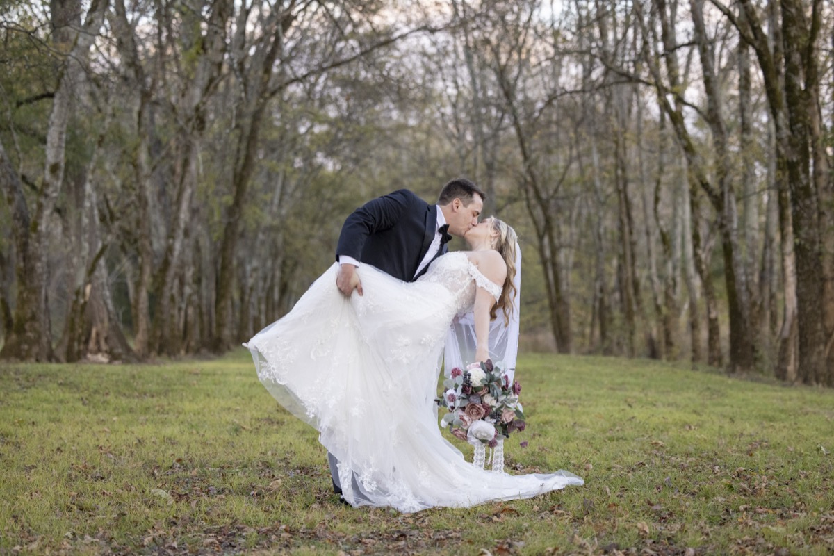 Kristen & Dalton dip-kiss in a grassy tree-lined field, burgundy bouquet in her hand at Covey Creek