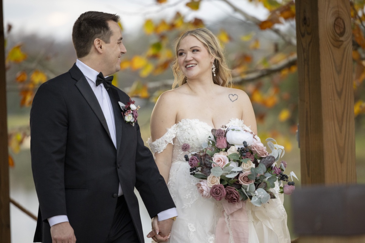 Kristen & Dalton walk hand-in-hand smiling, mauve bouquet and black tuxedo under autumn trees at Covey Creek