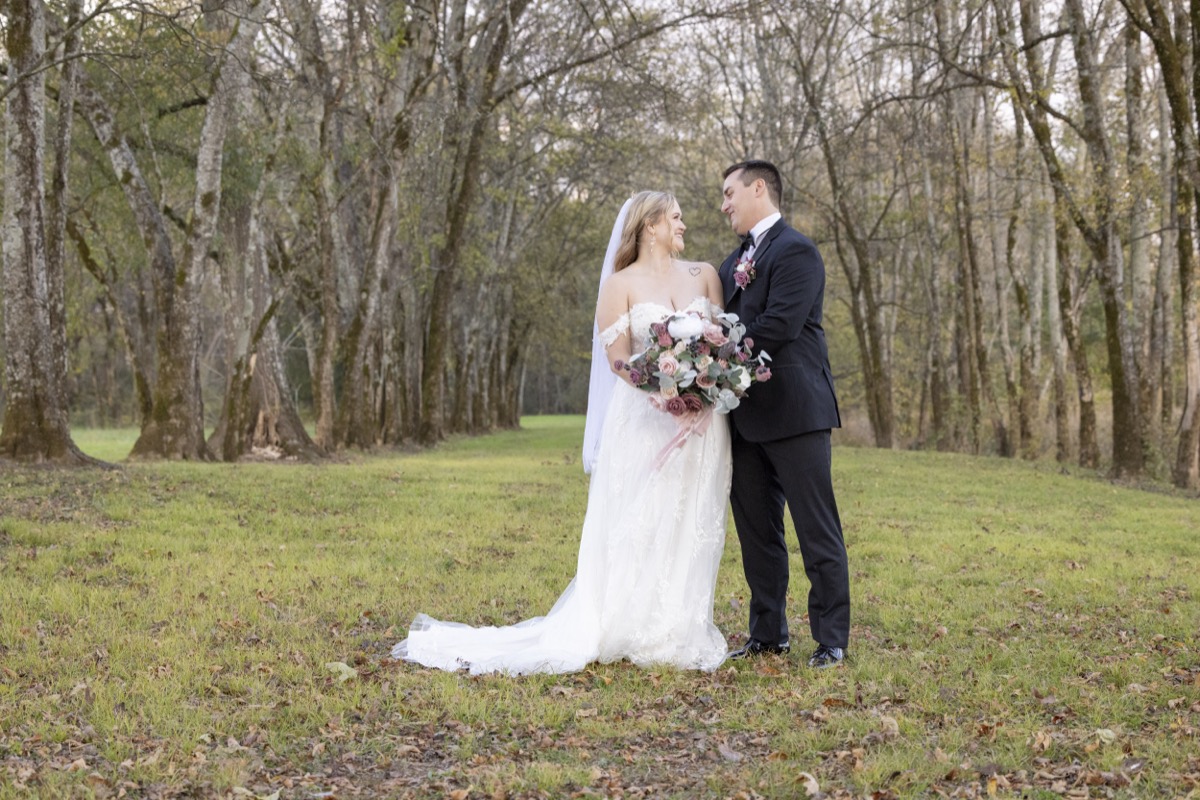 Kristen & Dalton smile face-to-face in a grassy field of tall trees, lace gown and black tuxedo at Covey Creek