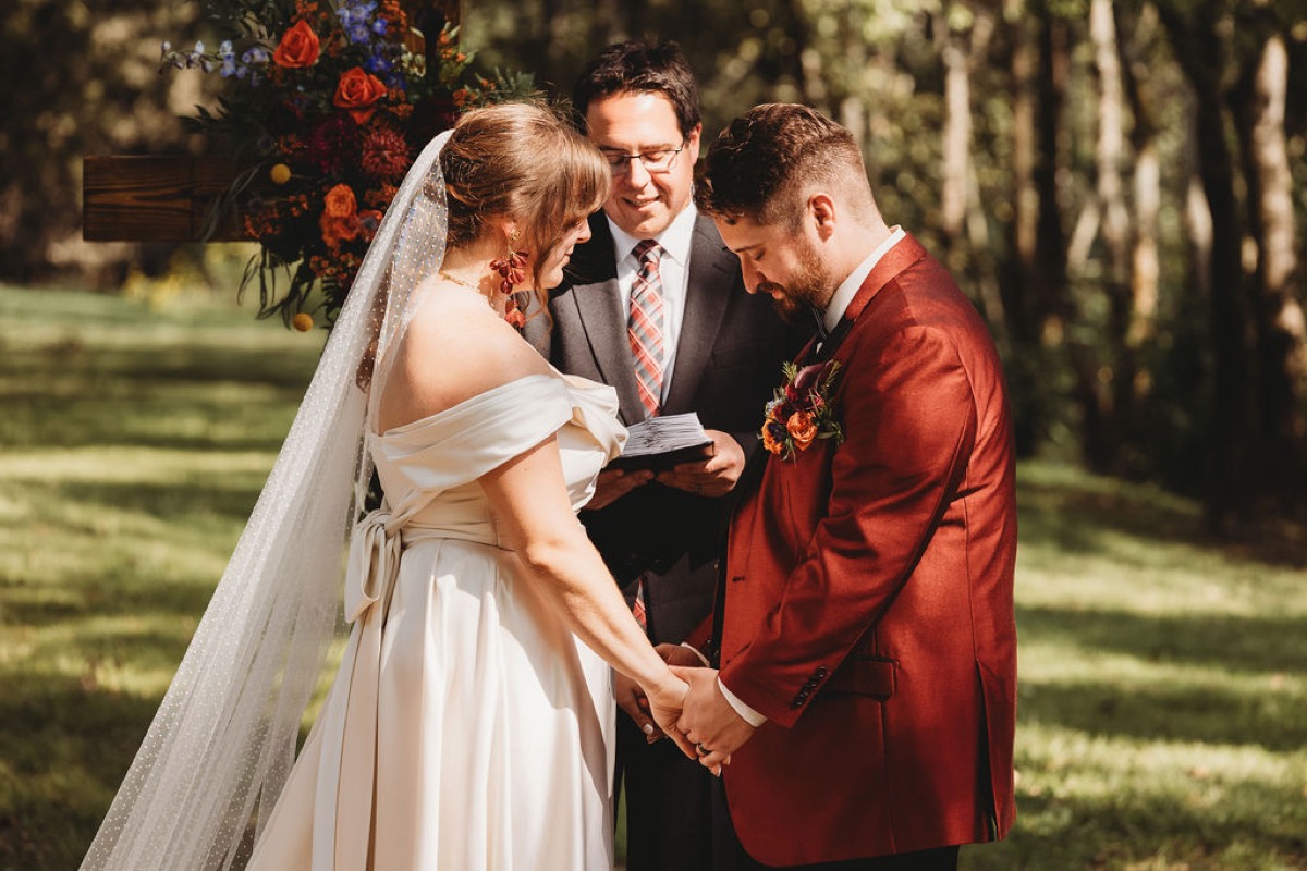Lauren & Alex bow their heads holding hands during outdoor vows by a floral cross at Covey Creek