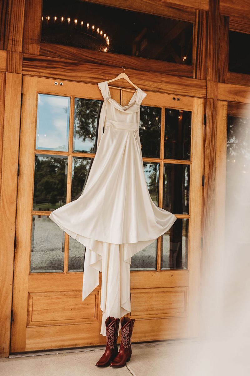 Lauren of Lauren & Alex's gown hangs on wooden doors above embroidered cowboy boots at Covey Creek