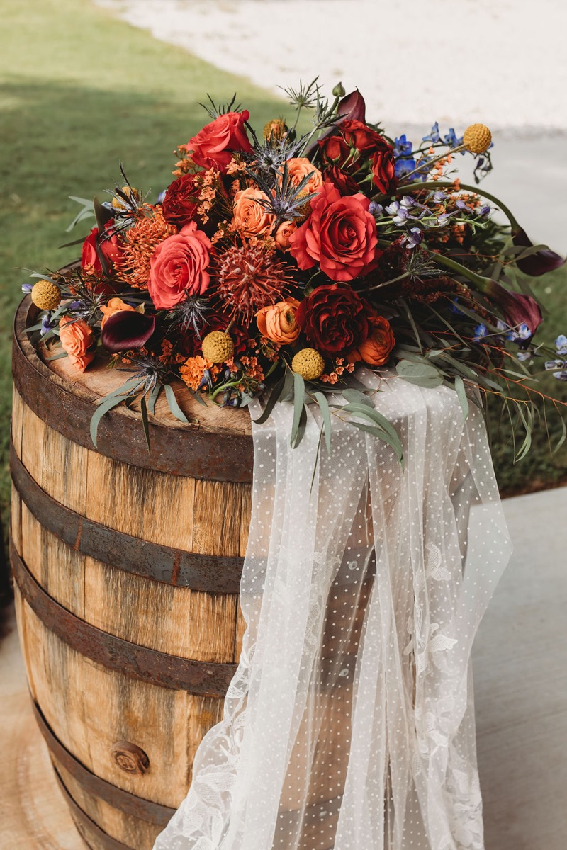 Autumn florals in red, orange, and blue sit on a wooden barrel for Lauren & Alex at Covey Creek