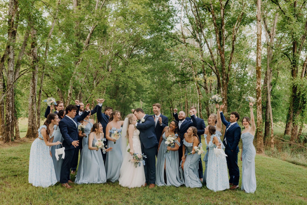 Leah & Gordon kiss as their bridal party cheers on Covey Creek’s green lawn against summer woods
