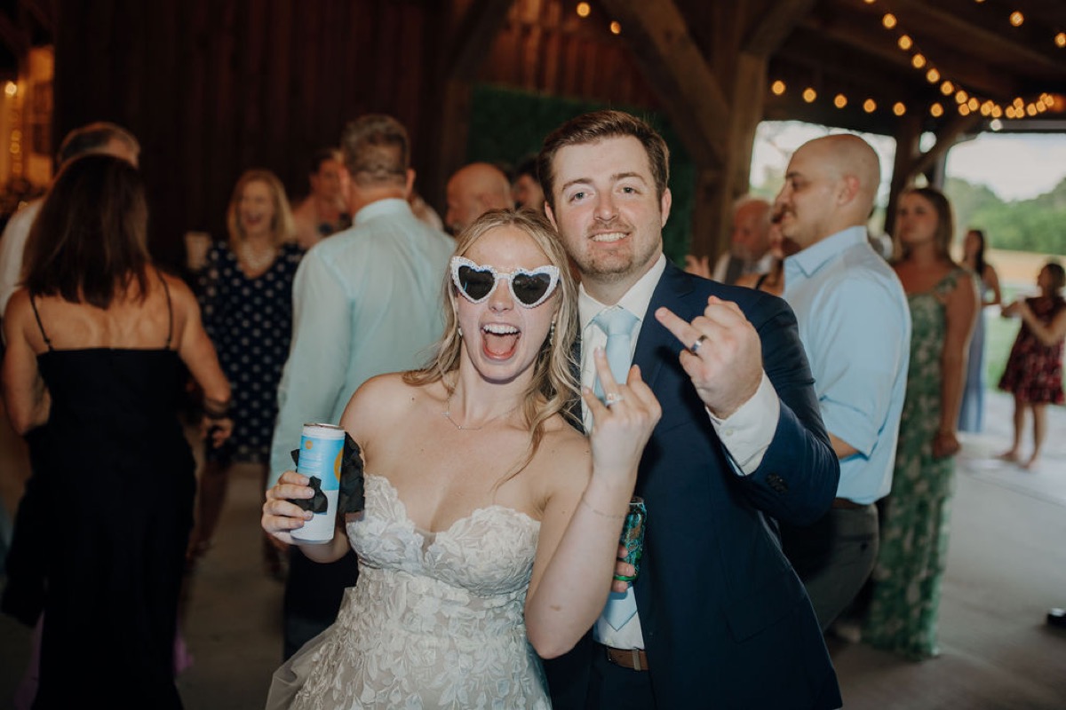 Leah & Gordon flash wedding rings in Covey Creek’s barn, heart sunglasses and navy suit mid-celebration