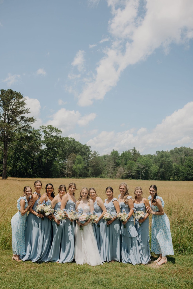 Leah & Gordon’s bridal party poses in a golden field at Covey Creek under blue sky and tree line