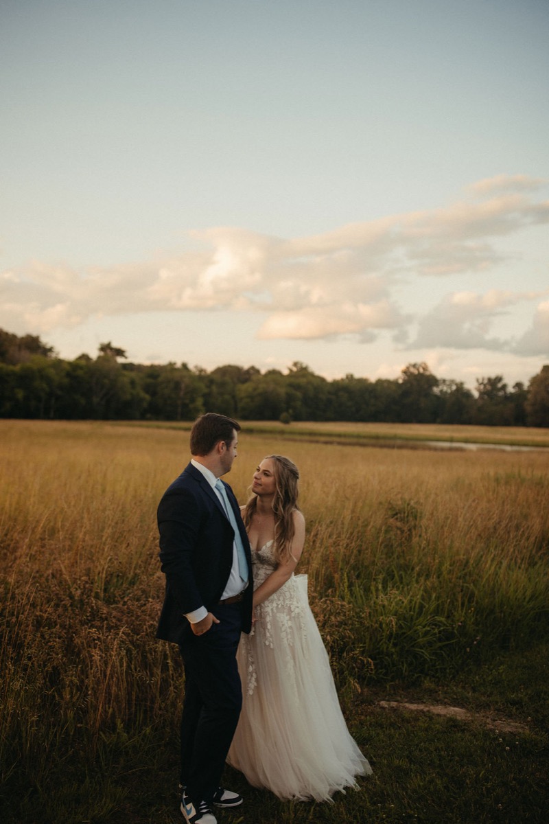 Leah & Gordon stand close in tall golden grass at Covey Creek, navy suit and lace gown at sunset