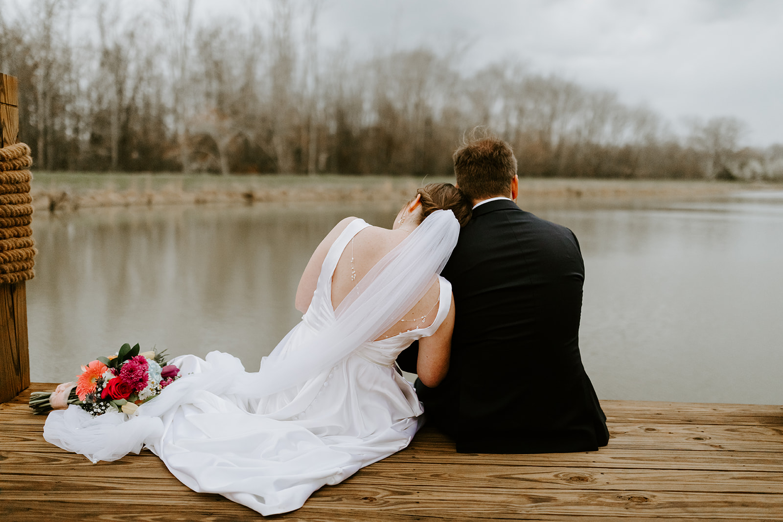 Leslie & Timothy sit close on the wooden pond dock with bouquet beside them at Covey Creek