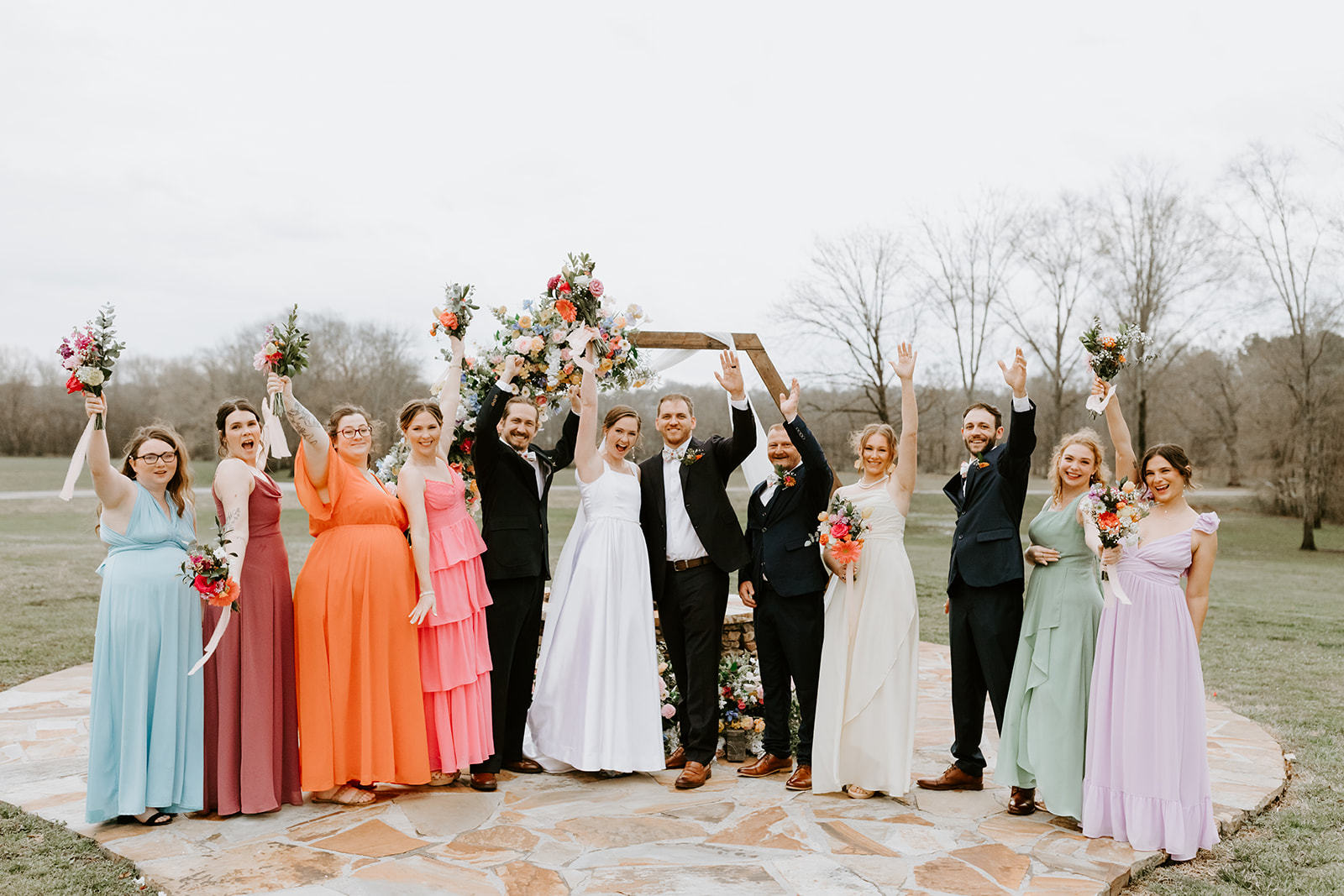 Leslie & Timothy raise arms with colorful wedding party before floral hex arch at Covey Creek