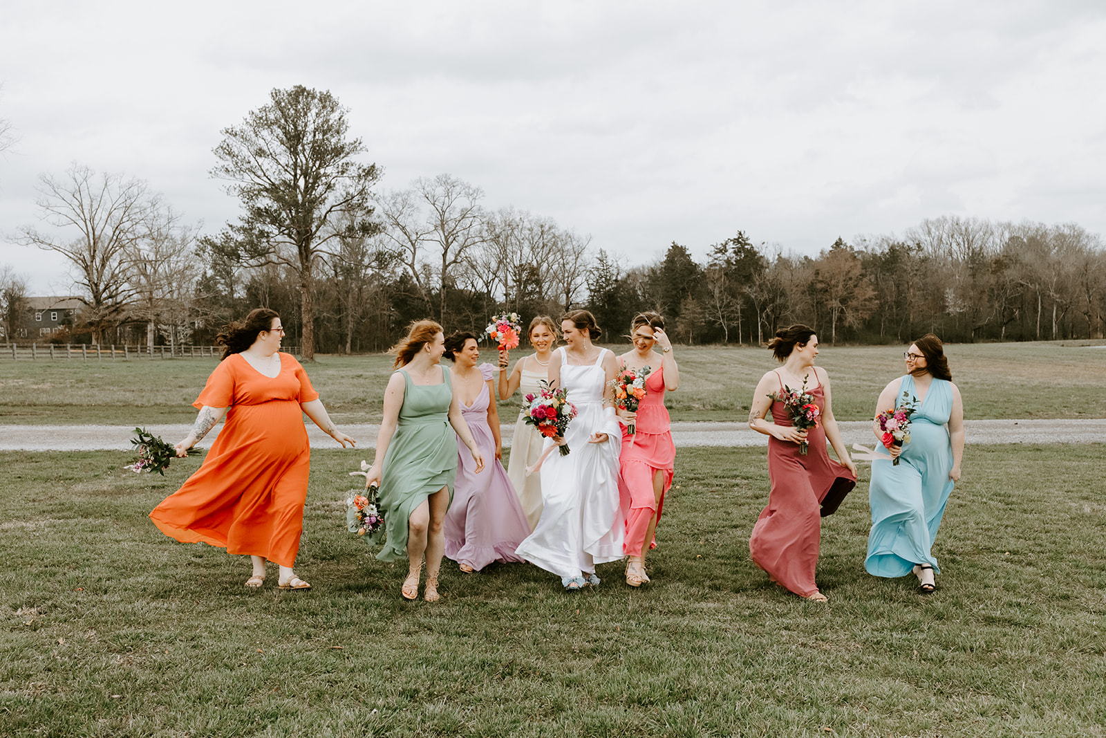 Leslie & Timothy bridal party walks laughing in rainbow dresses across a Covey Creek field