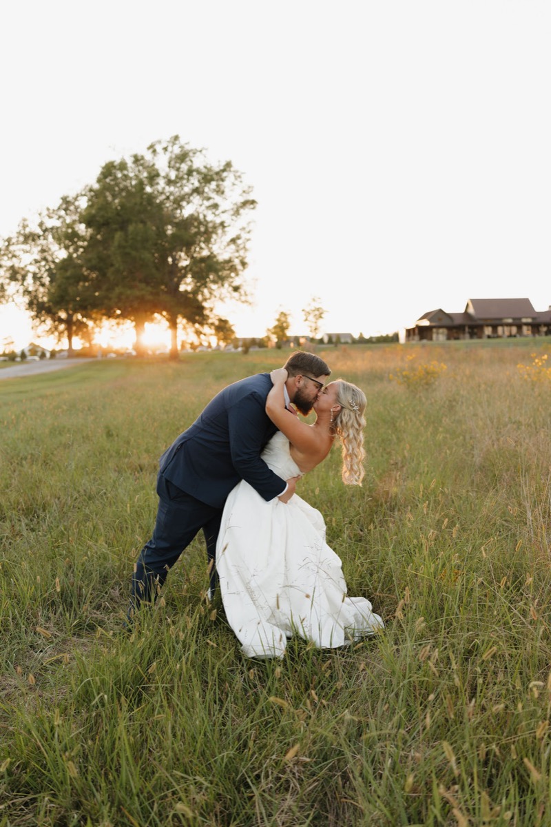 Lilly & Jackson share a golden-hour dip kiss in tall grass with the Covey Creek venue behind them at sunset