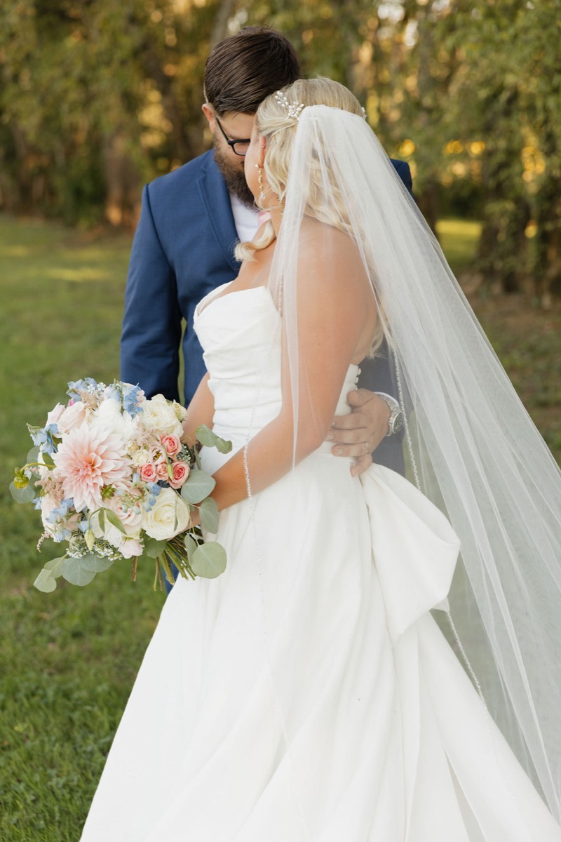 Lilly in veil holds a peach-pink bouquet while embracing Jackson outdoors at Covey Creek for Lilly & Jackson